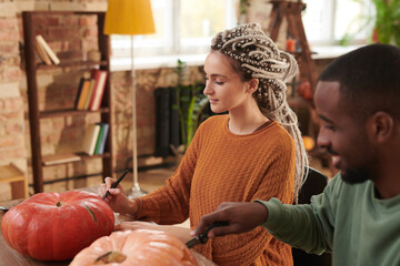 Concentrated young woman with dreads sitting at table with Black guy and cutting into pumpkins while making jack-o-lantern for Halloween
