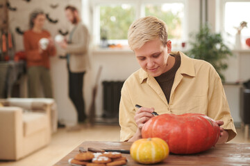 Content young blond woman in yellow shirt sitting at table and drawing face design on pumpkin while making Halloween decoration