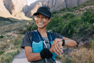Smiling woman hiker in cap looking away. Female trail runner checking smart watch while standing in...