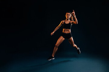 Young woman runner jumping in studio. Muscular athlete exercising on black backdrop.