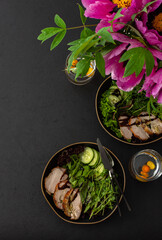 Hearty delicious lunch, fresh salad with cucumbers and microgreens of peas and with stewed pork, top view of the dark table with food and a vase of flowers