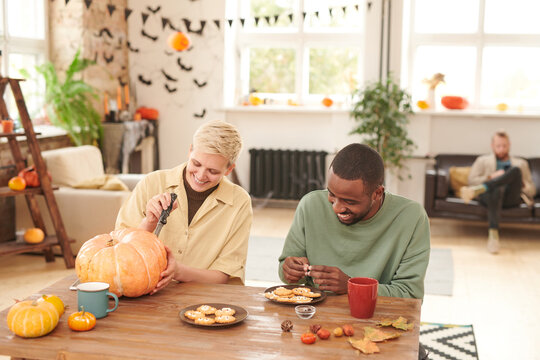 Positive young multi-ethnic friends sitting at wooden table and enjoying Halloween preparation: Black man decorating cookies while blond girl carving pumpkin