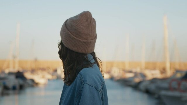 Young beautiful woman standing on yacht club pier on warm sunset, dreaming, thinking, turning around and smiling gently at camera. Pretty female wearing cosy casual cloths on trip, city exploration
