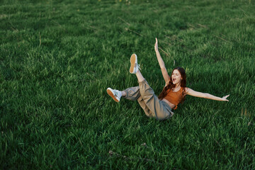 A young woman playing games in the park on the green grass spreading her arms and legs in different directions falling and smiling in the summer sunlight