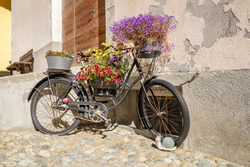 Fototapeta na wymiar An old rusty bike, decorated with beautiful flowers, in the historic village center of S-Chanf (Upper Engadine Valley , Grisons, Switzerland). It probably hasn't been ridden in a long time.