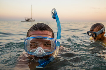 Portrait of a young man swimming in the sea from his yacht at sunset snorkeling with his little son who is also wearing a swimming mask. - obrazy, fototapety, plakaty