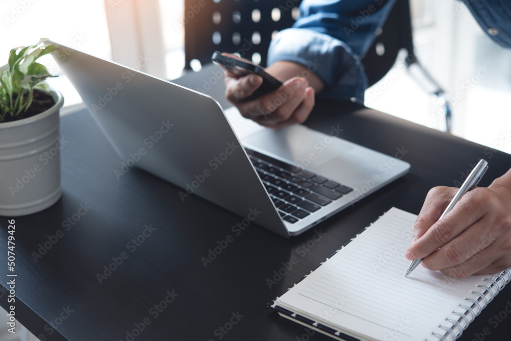 Poster closeup of man hand with a pen writing on notebook, working on laptop computer and using mobile smar