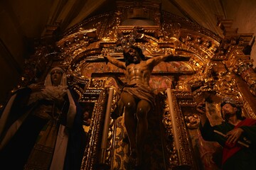 Sculpture of a very expressive Jesus at the cross wearing shiny plateresque crown, brilliant golden Baroque ornaments in the back at Santa María de la Asunción basilica, Arcos de la Frontera, Spain