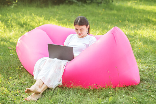 Adult Woman Working Or Studying Remotely Using Laptop And Internet. Summer. Pink Bean Bag Chair.