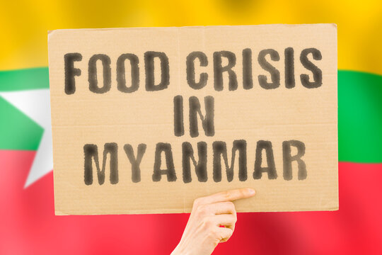 The phrase " Food crisis in Maynmar " is on a banner in men's hands with a blurred Myanmarese flag in the background. Crisis. Finance. Life. Nutrition. Bread. Disaster. Collapse. Social issue