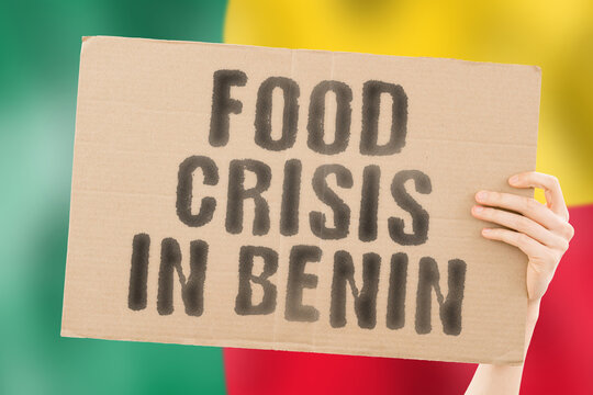 The phrase " Food crisis in Benin " is on a banner in men's hands with a blurred Beninian flag in the background. Crisis. Finance. Life. Nutrition. Bread. Disaster. Collapse. Social issue