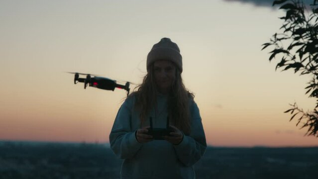 Soft sunset twilight view of young female holding, using joystick to control copter, flying drone outside on stunning natural background to take shots, videos for travel hobby blog, explorer website