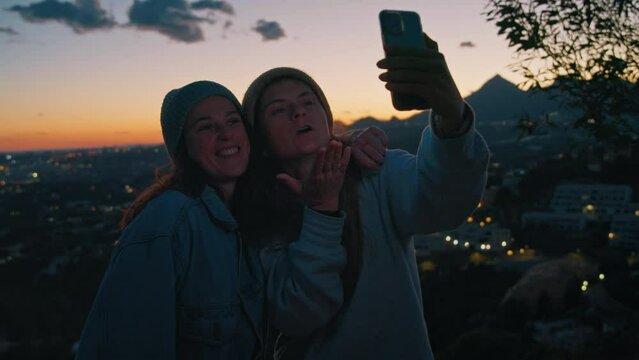 Young millennial cheerful women takes happy selfie, chats by video with friends, sending kisses, flirting, using frontal camera on smartphone, posing on outdoor camping trip near sea, ocean on sunset