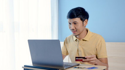 Handsome Asian man, wearing a yellow shirt, looks happy when he successfully pays for an online purchase via his laptop computer with a credit card,online shopping.
