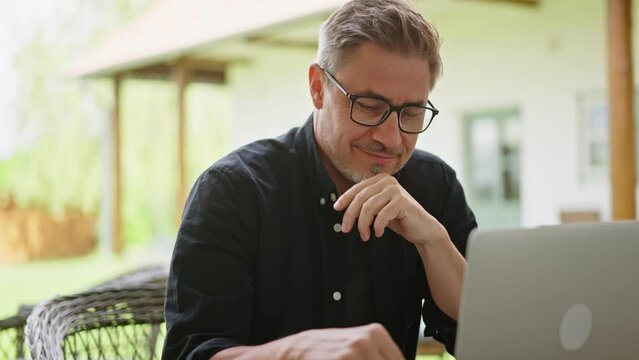 Casual middle age man sitting in garden working on laptop computer in home office. Businessman tele working