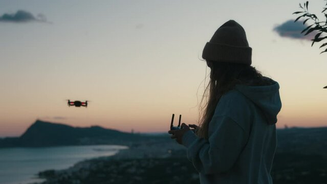 Soft view of young female silhouette holding, using joystick to control copter, flying drone outside on stunning sunset windy natural background to take pictures, videos for travel hobby blog, website