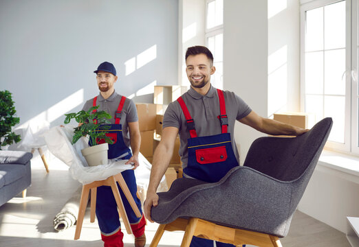 Team of happy workers from truck delivery and moving service company together removing furniture and other stuff from apartment. Two strong men in uniforms carrying chairs, armchairs and house plants