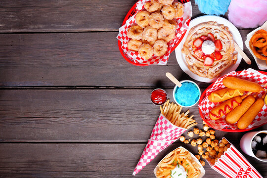 Carnival theme food side border over a dark wood background. Top view with copy space. Summer fair concept. Corn dogs, funnel cake, cotton candy and snacks.
