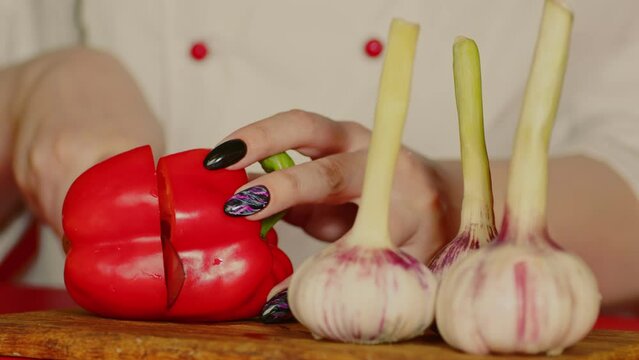 Red sweet pepper with young garlic on wooden board. Unrecognizable woman cuts red bell pepper with knife.