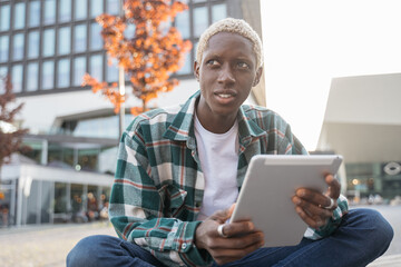 Portrait of handsome smiling African American student using digital tablet studying, learning language sitting in university campus, online education concept. Stylish freelancer working outdoors  