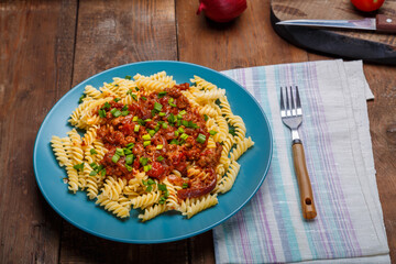 A blue plate with bolognese pasta on a wooden table on a light napkin next to a fork.