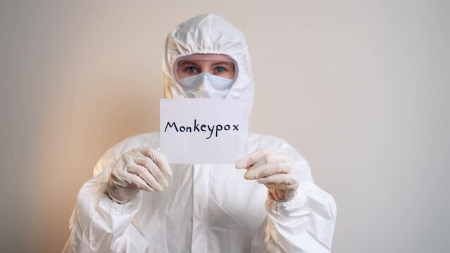 Isolated woman in protective suit, mask and goggles holding a sheet of paper with the inscription monkeypox. Medium plan. A new virus in medicine. Infectious disease. A new kind of pandemic.
