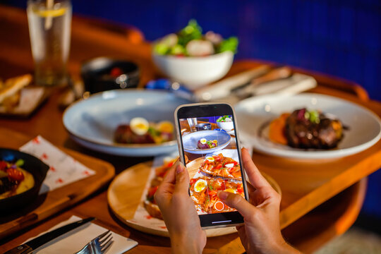 Woman photographing salmon toast through mobile phone at table in restaurant. Hands hold smartphone taking photo of fine dining food in cafe. Smartphone food photography of lunch or dinner