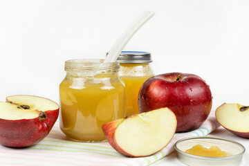 Baby food. Two glass jars with baby apple puree with a spoon and lid, fresh red apples on a white background. The first fruit lure of the baby.
