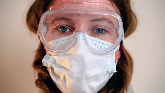 Female doctor during coronavirus pandemic covid-19 takes off glasses, hood from protective suit and mask, face marks are visible, red spots. Nurse inhales and exhales air with relief.Close up portrait