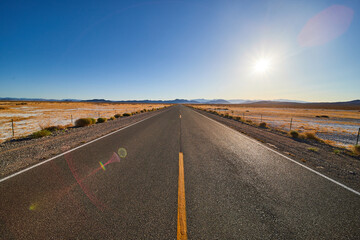 Desert paved road leading into endless desert with sun and blue sky