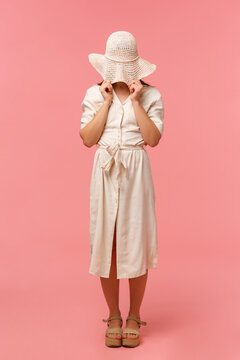 Full-length Vertical Portrait Silly And Shy Woman Standing In Dress, Hiding Her Face Behind Summer Hat, Standing Over Pink Background, Embarassed Look, Feeling Awkward Or Blushing