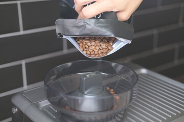 A hand pours coffee beans from a black bag into a coffee machine against a black wall in the kitchen