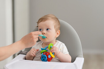 Baby boy eating blend mashed food sitting, on high chair, mother feeding child, hand with spoon for vegetable lunch, baby weaning, first solid food for young kid.