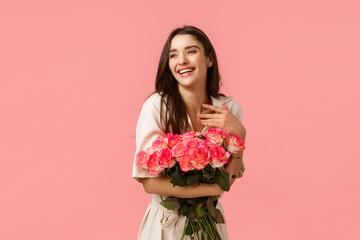 Happiness, romance and beauty concept. Cheerful gorgeous brunette girl in dress, holding roses and smiling flattered, touch chest laughing look away blushing, standing pink background enthusiastic