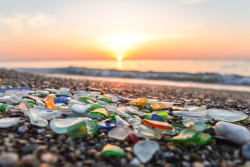 Colored sea glass with beach pebbles and shells in the mediterranean coast and in the background sea and waves with sunset and sky, sandy beach with coast glass, beach glass. - obrazy, fototapety, plakaty