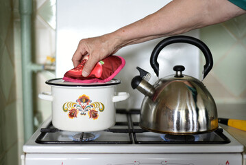 The hand of an elderly woman lifts the lid of a saucepan standing on a gas stove.