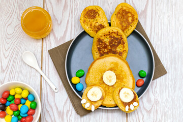 Easter funny creative healthy breakfast lunch food idea for kids, children.Bunny, rabbit made from pancakes,banana with sweet candies,honey on plate wooden table background.Top view Flat lay