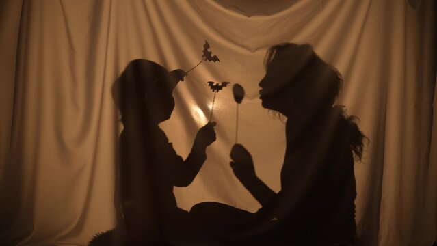 Unrecognizable woman and boy telling spooky stories using shadow play and paper bats and pumpkins. Mom and son have fun, sitting behind the curtain at home. Puppet theater of shadows. Close up.
