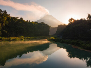 Lake with a water surface that looks greenish and emits smoke in the morning with a volcano on the background in sunrise. The lake surrounded by trees. The volcano named Merapi Volcano in Indonesia.