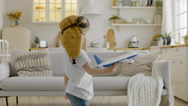 Child aspires to fly high in clouds playing with an airplane at home. Happy child in aviapilot hat is holding toy airplane in his hands, circling with an airplane and dreaming of flying in clouds.