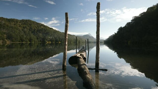 Magical natural scenery. View of a rustic dock made with tree trunks and branches, in lake Espejo Chico in Patagonia. The green forest, Andes mountains and sky reflect in the water surface.