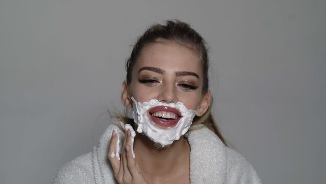 Attractive young woman shaves face. Cheerful funny woman has having cream over her face. She is shaving herself with razor.