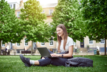 A young girl smiles looking at her laptop. A woman works in a laptop sitting on the grass. A woman takes online courses in the park, sitting on the lawn. The concept of a remote office.