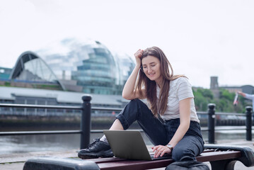 A cheerful girl works with a laptop on the embankment sitting on a bench. The concept of an open-air office. Freelancer at work. Newcastle, The Sage Gateshead, North East England. - obrazy, fototapety, plakaty