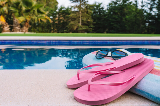 close up of summery swimming objects in front of a swimming pool. beach towel, sunglasses, swimming flip-flops. lawn garden with swimming pool in the background.