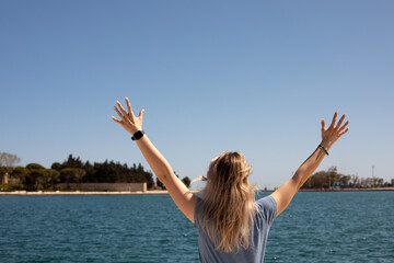 Rearview of blond woman stand against blue sky with raised arms embracing world, sea vacation and...