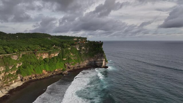 Aerial drone footage of Pura Luhur Uluwatu on sunset, one of Bali's most famous sea temples, perched on dramatic limestone cliffs overlooking the Indian Ocean. Waves crash against rugged coastline.