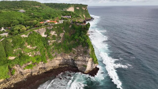 Aerial drone footage of Pura Luhur Uluwatu on sunset, one of Bali's most famous sea temples, perched on dramatic limestone cliffs overlooking the Indian Ocean. Waves crash against rugged coastline.