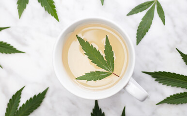 white mug with marijuana infusion with green leaf inside on marble table with leaves. top view.