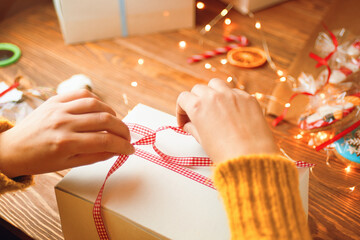 Woman's hands in knitted sweater tie red ribbon bow on white gift box. New Year's mood. Garland lights, gingerbread and decorations in background. Surprise for Birthday or Christmas.